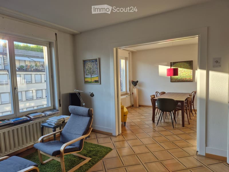 Bright living and dining area with wooden flooring, large windows, a blue armchair, a small table with books, a dining table with chairs, and artwork on the walls.