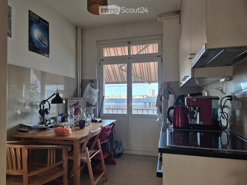 A modern kitchen with a wooden dining table, chairs, a coffee maker, kettle, and a lamp on the counter. There is a view of the sea through the windows.