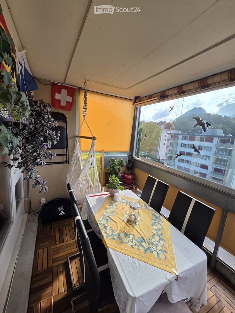 Balcony with wooden floor, large window offering a scenic view, table with chairs, hanging chair, plants, and decorative items.