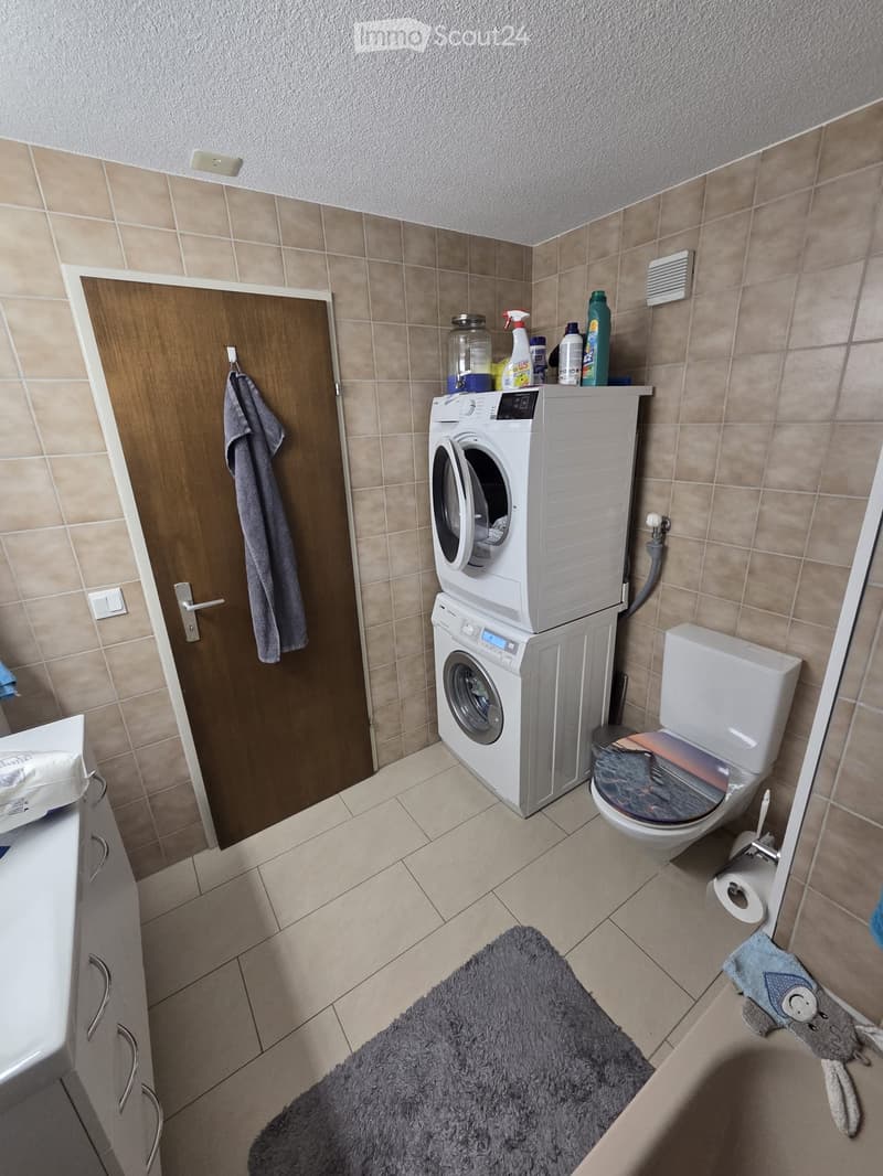 Bathroom with tiled walls and floor, featuring a stacked washing machine and dryer, toilet, cabinet, and cleaning supplies on top of the dryer. A towel hangs on the door, and a rug is placed on the floor.
