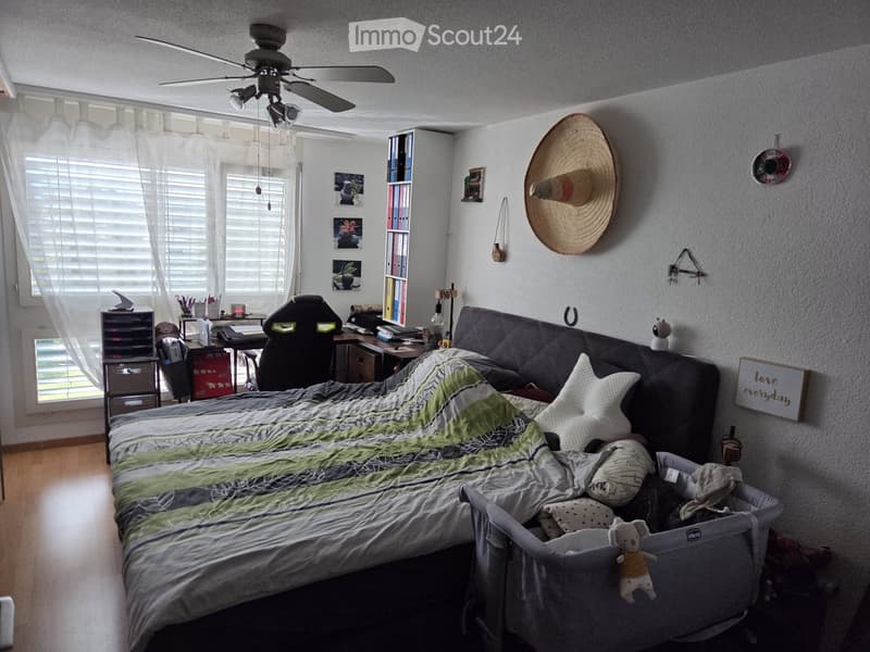 Cozy bedroom with a large bed, a sofa, a baby crib, a desk, and various decorative items including a large hat on the wall and framed pictures.