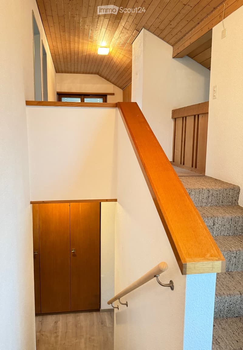 Wooden staircase with handrail and wooden door in the background, white walls, and wooden ceiling.