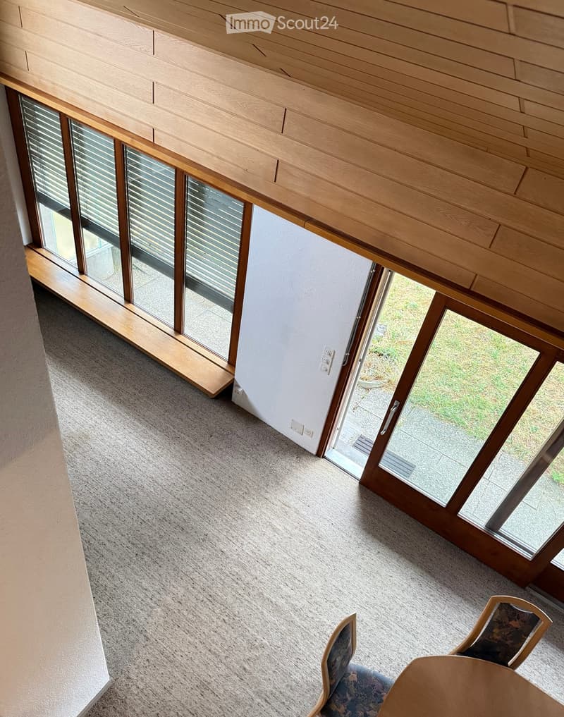A modern, minimalist room with large windows and wooden sliding doors leading to an outdoor area. The room features a light-colored carpet, a wooden ceiling, and a small seating area with a table and chairs.