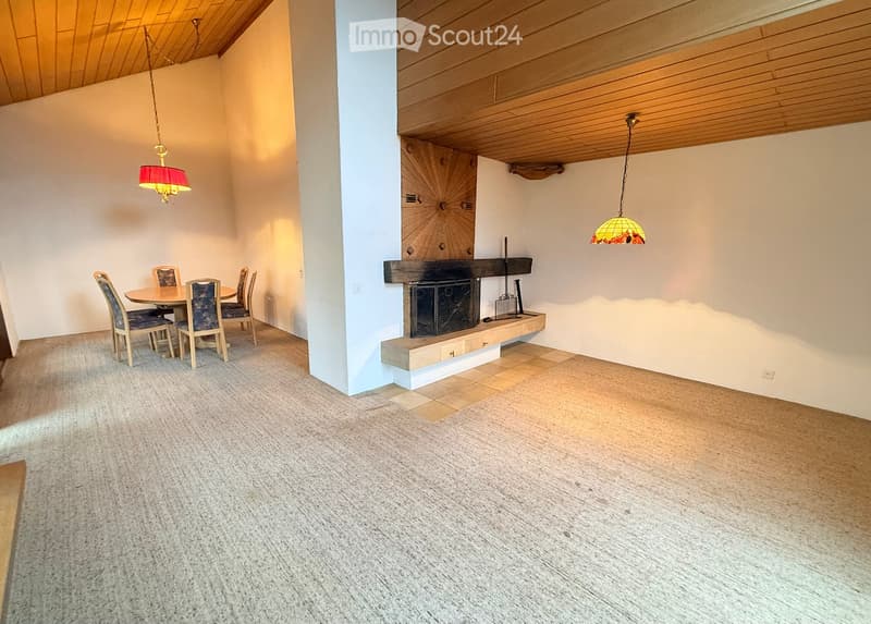 Dining area with a round wooden table, four chairs, and a fireplace with a wooden mantle. The room features a mix of carpet and wood flooring, and two hanging lamps provide lighting.