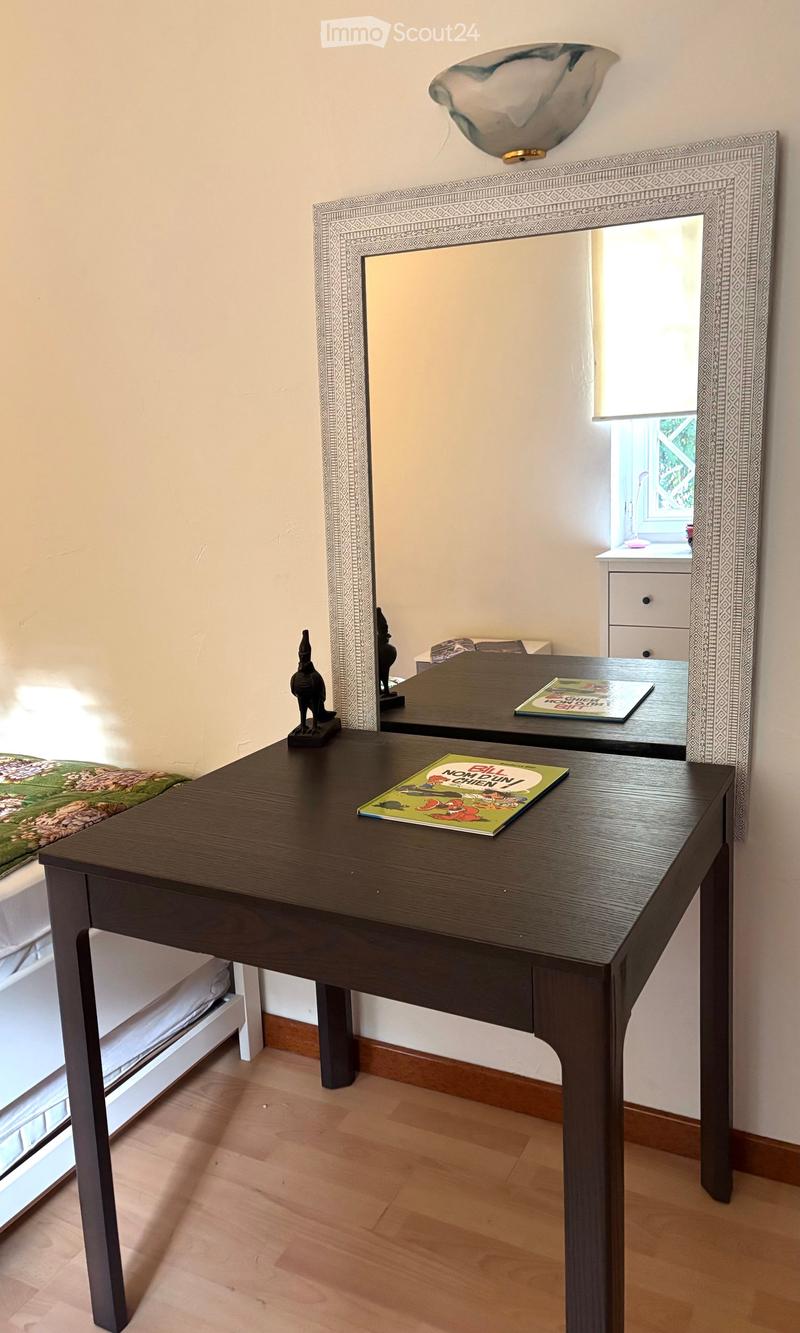 Desk with a book, mirror on the wall, bed, and a reflection of a room