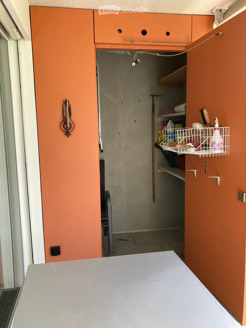 A small utility or storage room with orange cabinets, a white table, shelves holding cleaning supplies, and a broom leaning against the wall.