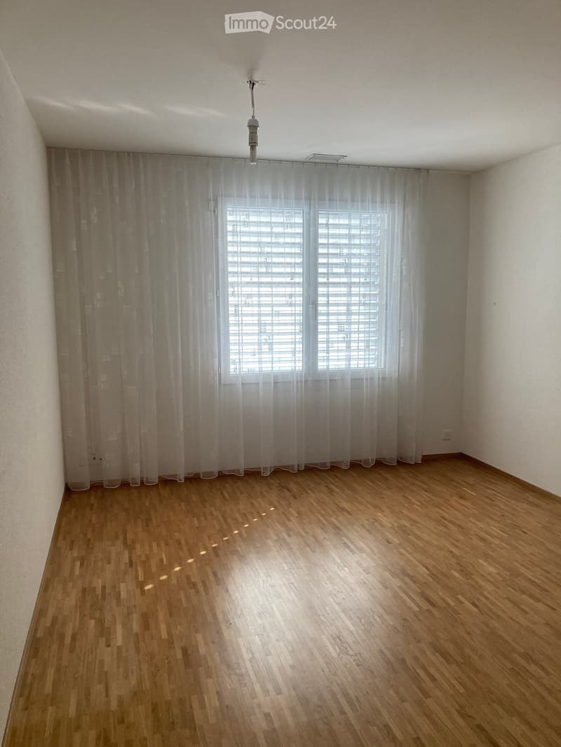 Empty room with wooden flooring, white walls, large window with blinds and sheer curtains, and a ceiling light fixture.