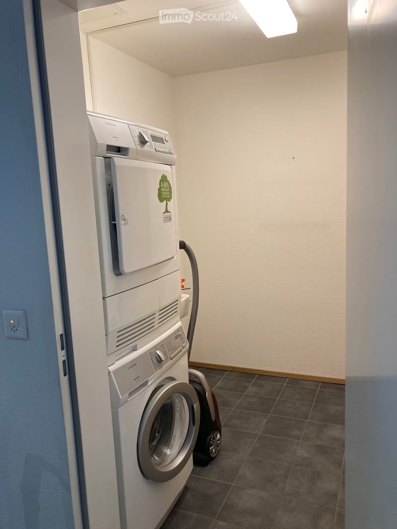 A small, well-lit room with a washing machine and a dishwasher stacked vertically, along with a vacuum cleaner on the floor.