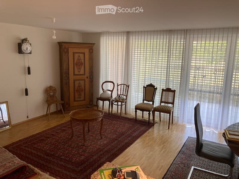 Elegant living room with wooden flooring, large windows with sheer curtains, a round wooden table, four chairs, a cabinet with framed artwork, a wall-mounted clock, and a mirror on the left side.