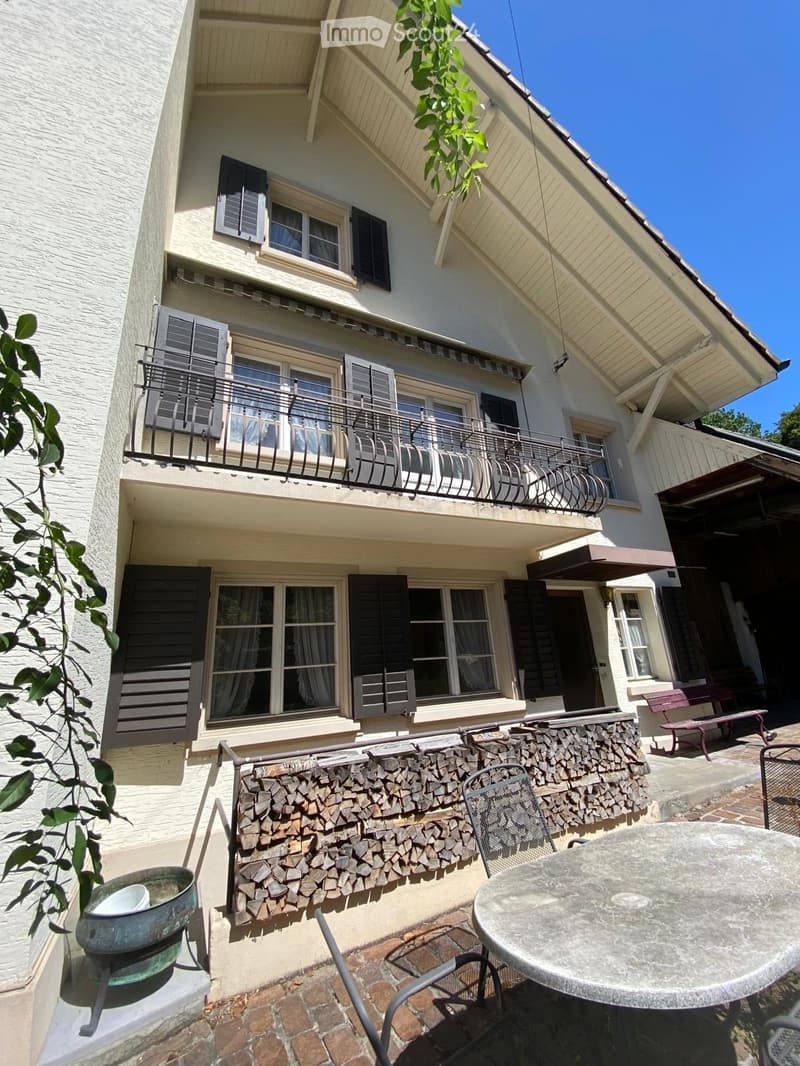 Outdoor patio area with a round table, chairs, a bench, and a stack of firewood against the wall of a two-story house.