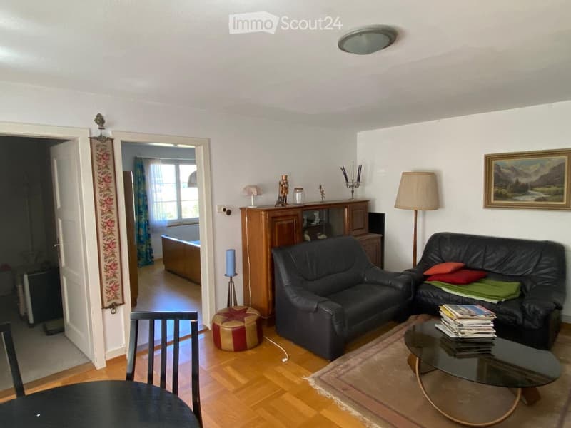A well-lit living room with wooden flooring, two black leather sofas, a glass coffee table, a wooden cabinet, and a decorative stool.
