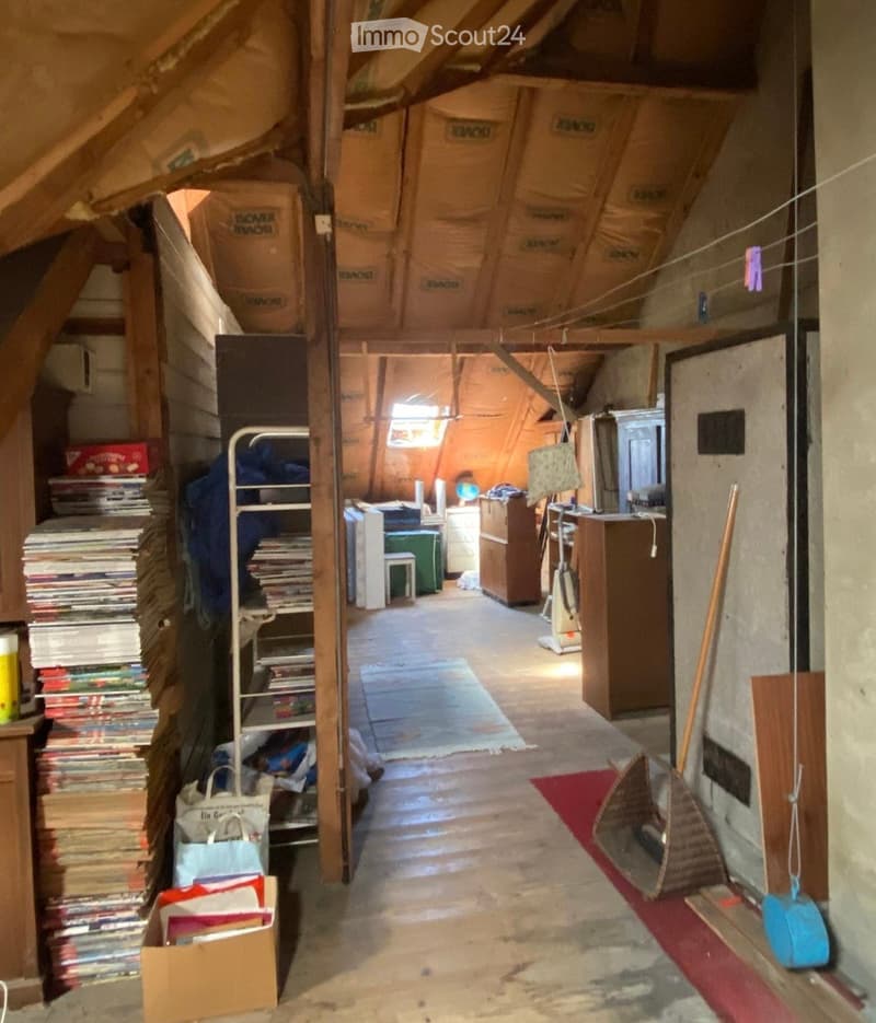 An attic space with wooden beams, exposed insulation, and various stored items including shelves of books, a small table and chair, a refrigerator, and a vacuum cleaner. The floor is concrete, and there are bags and boxes scattered around.
