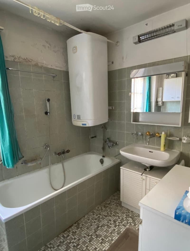 Bathroom with a bathtub, sink, mirror, and water heater. Green tiles on walls and floor, with a shower hose attached to the bathtub.