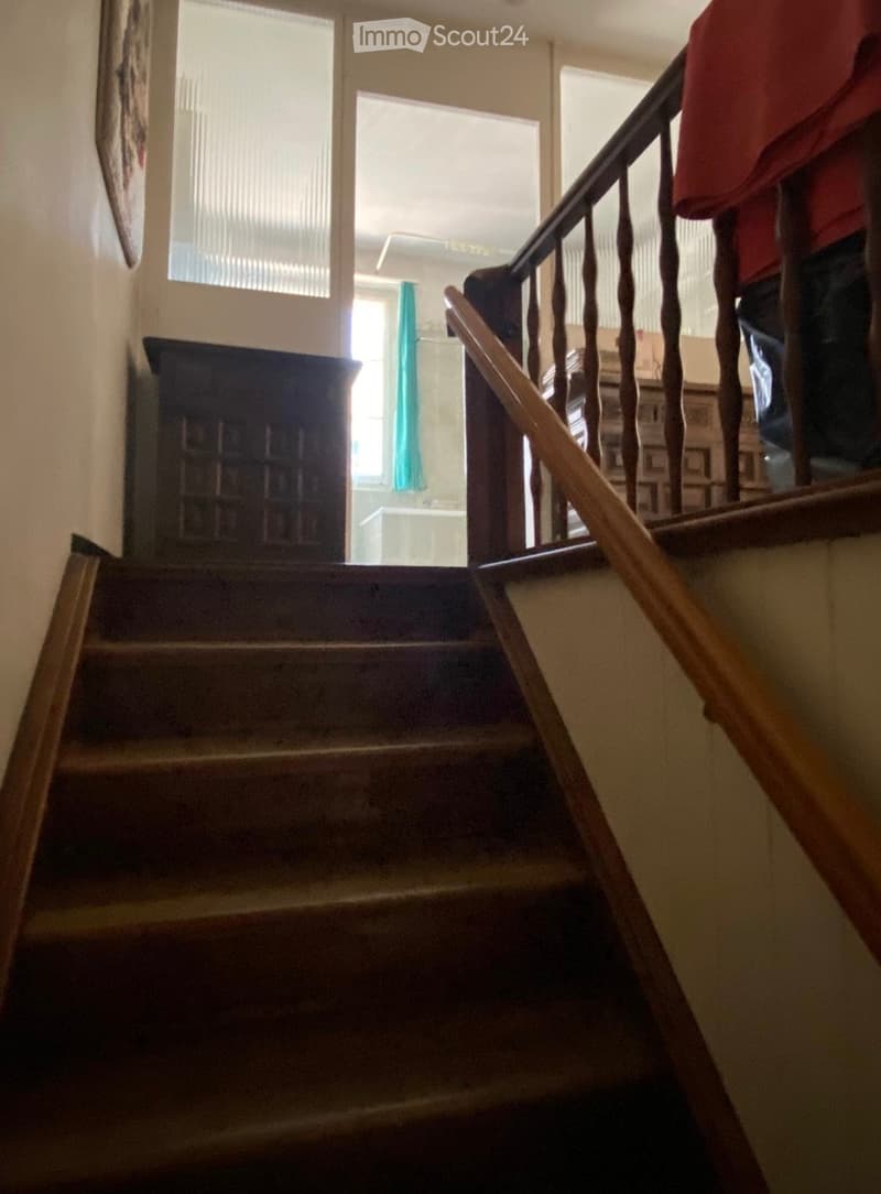 Wooden staircase with a wooden handrail and balusters, leading to an upper level with visible windows and a piece of furniture in the background.