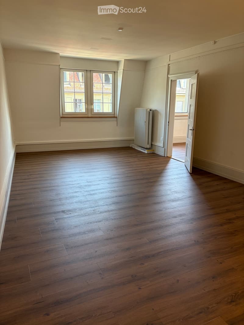 Empty room with wooden flooring, white walls, a radiator, and a window with a view of neighboring buildings.