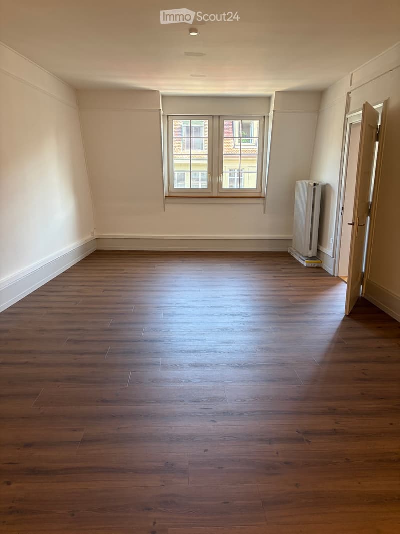 Empty room with wooden flooring, white walls, and a window with a view of a neighboring building. A radiator is visible on the right side.