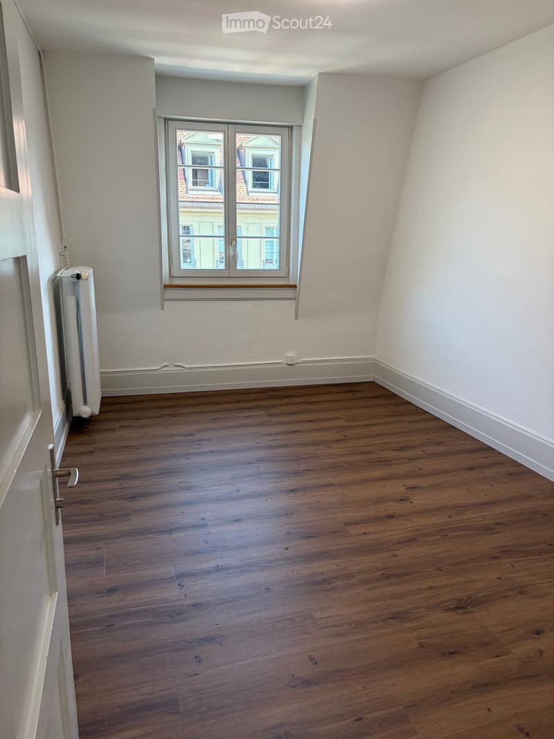 Empty room with wooden flooring, white walls, and a window with a view of neighboring buildings.