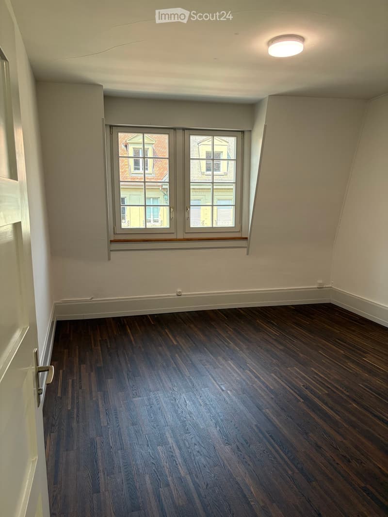 Empty room with wooden flooring, white walls, and a large window offering a view of neighboring buildings.