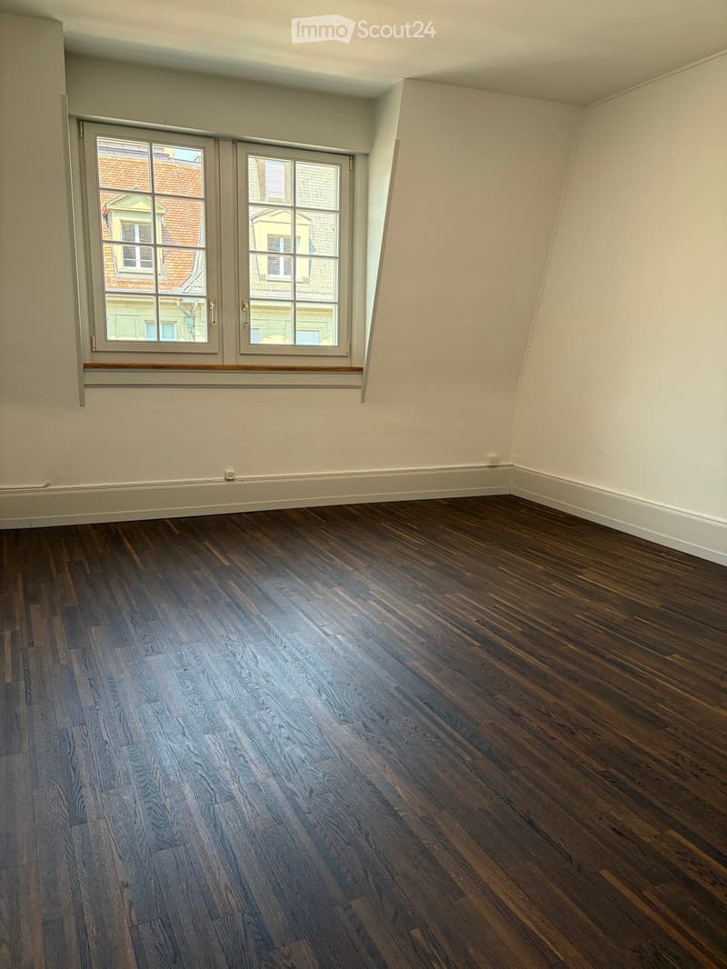 Empty room with wooden flooring and large windows showing a view of neighboring buildings.