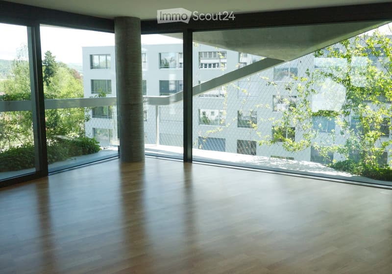An empty room with large windows and a polished wooden floor, offering a view of a modern building and greenery outside.