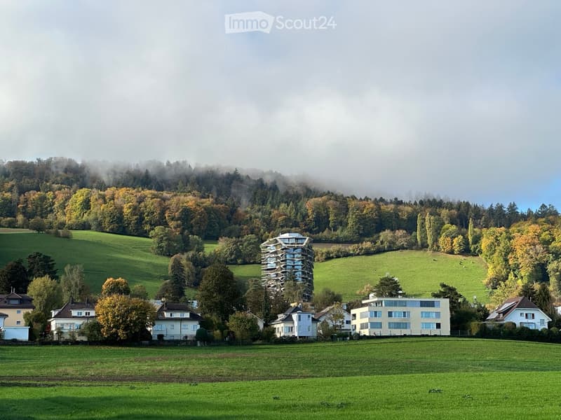 A modern, multi-story building with a unique, curved design stands prominently in a lush, green landscape surrounded by trees and rolling hills.