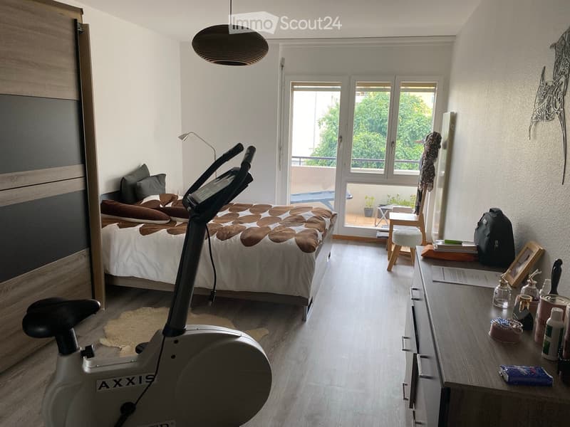 A modern bedroom with a large bed, exercise bike, nightstand, and a small table with various items. The room features large windows with a view of greenery and a balcony area.