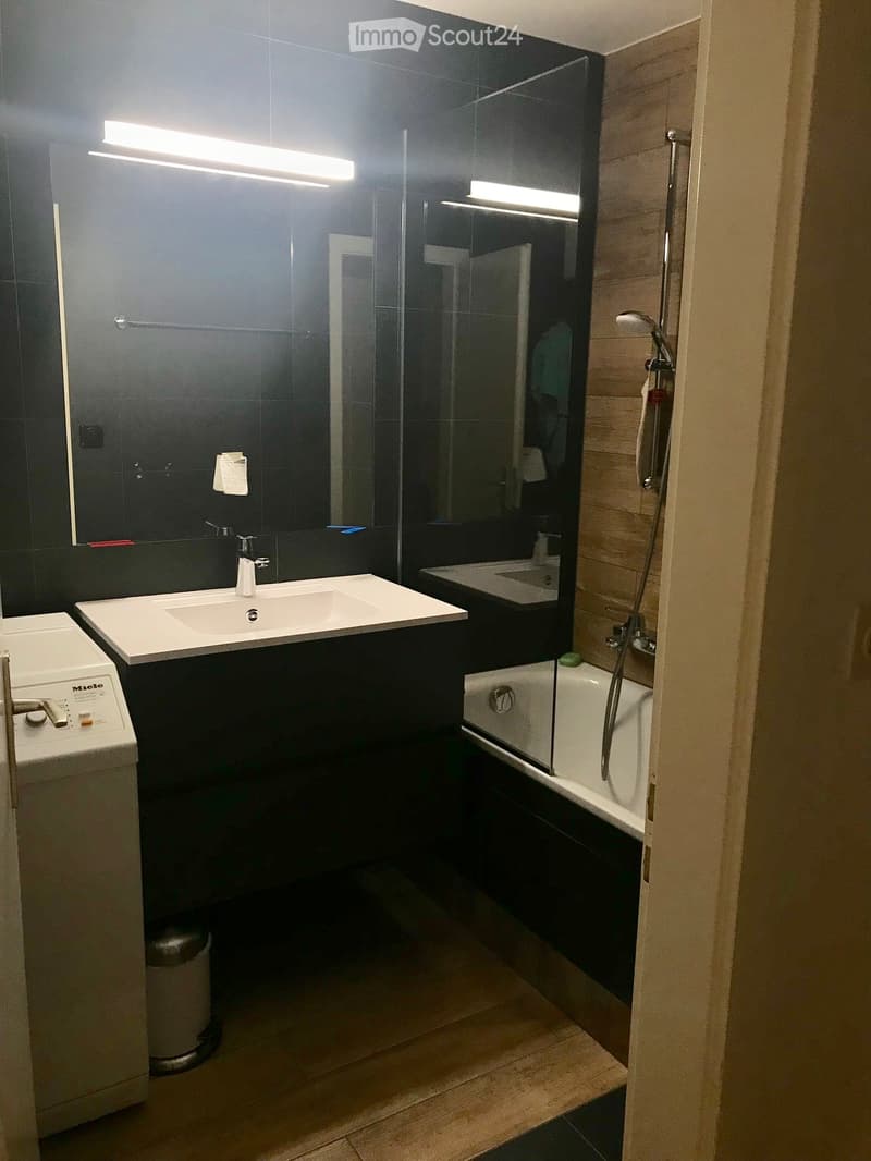 Modern bathroom with a white sink, bathtub with glass enclosure, shower, mirror, and a washing machine. Dark tiled walls and wooden flooring.