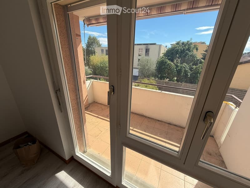 Open sliding glass doors lead to a tiled balcony with a metal railing, overlooking a residential area with trees and buildings.