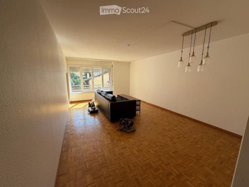 Living room with wooden herringbone flooring, white walls, large windows, and a black leather sofa.