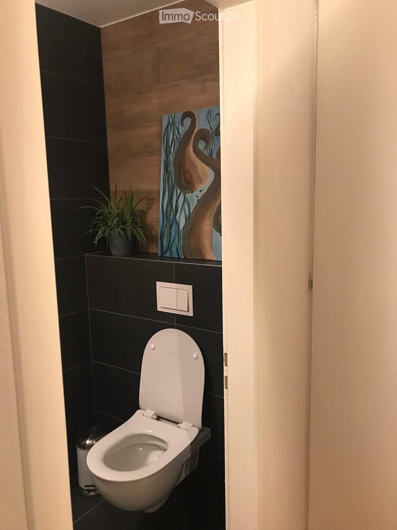 Modern bathroom with black tiled walls and floor, white toilet with open lid, small potted plant on shelf, decorative painting above, and light switch on wall.