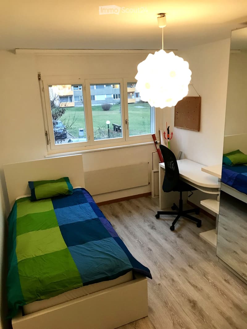 Cozy bedroom with a single bed, desk, chair, and a hanging lamp. The bed has a colorful quilt, and the room has large windows with a view of the outdoor area.
