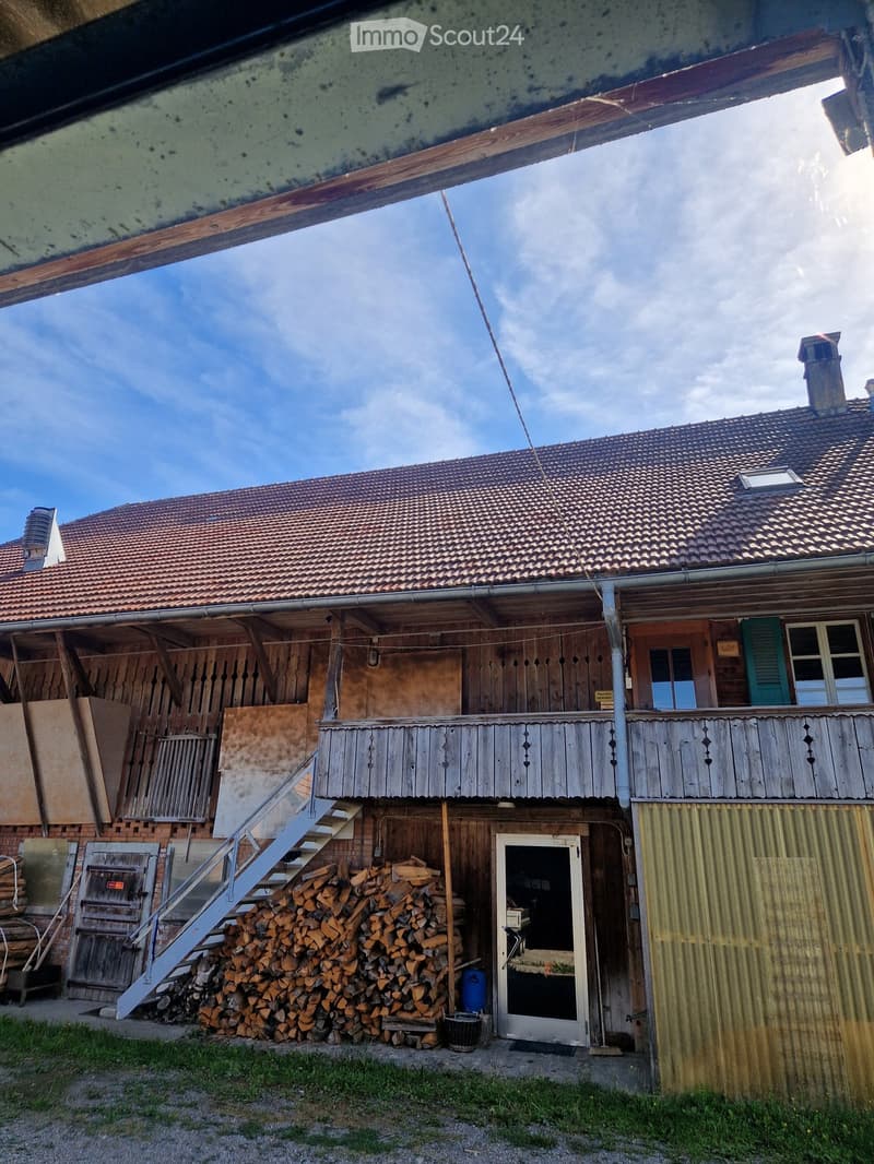 Two-story wooden house with a tiled roof, wooden balcony, and a stack of firewood in front.