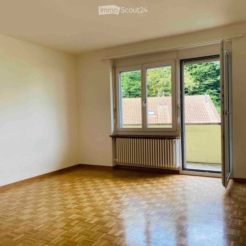 Empty room with wooden flooring, large windows, radiator, and a sliding door leading to a balcony.
