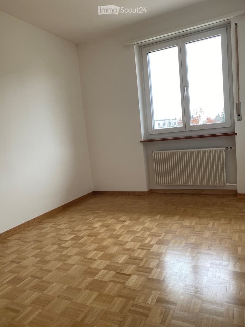 Empty room with wooden herringbone flooring, white walls, a large window with white frame, radiator, and a small shelf under the window.