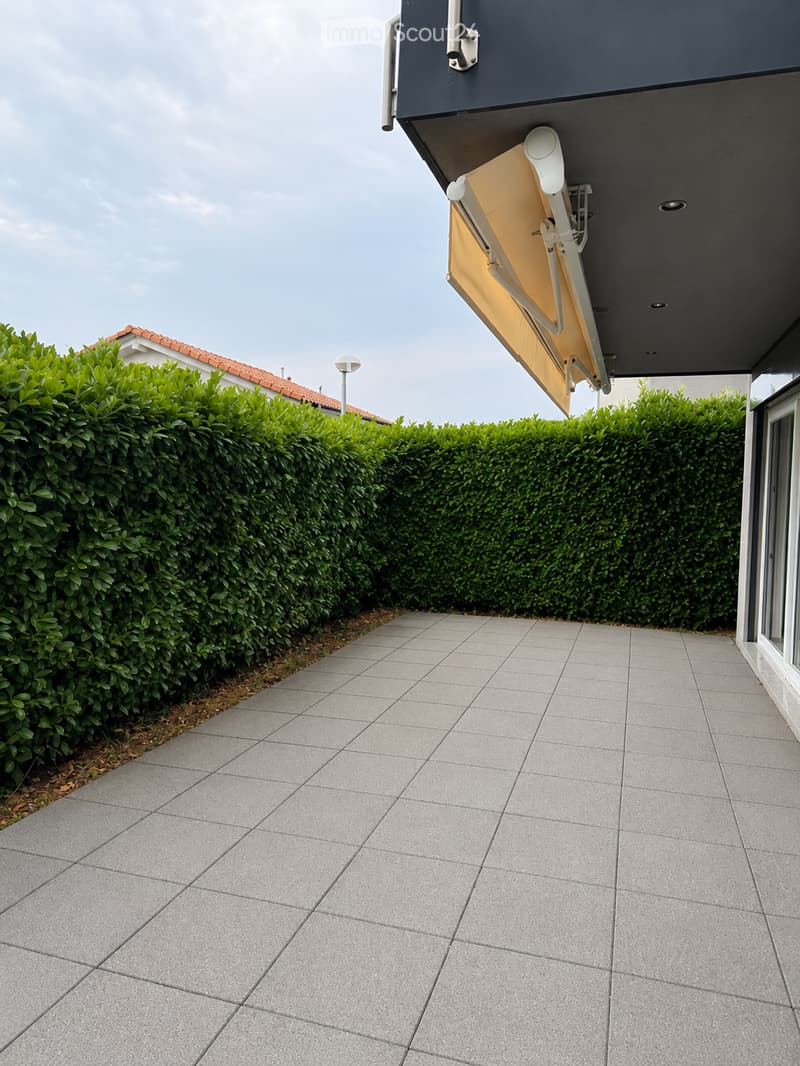 Grey tiled balcony with a neatly trimmed green hedge border and a yellow retractable awning attached to the building.
