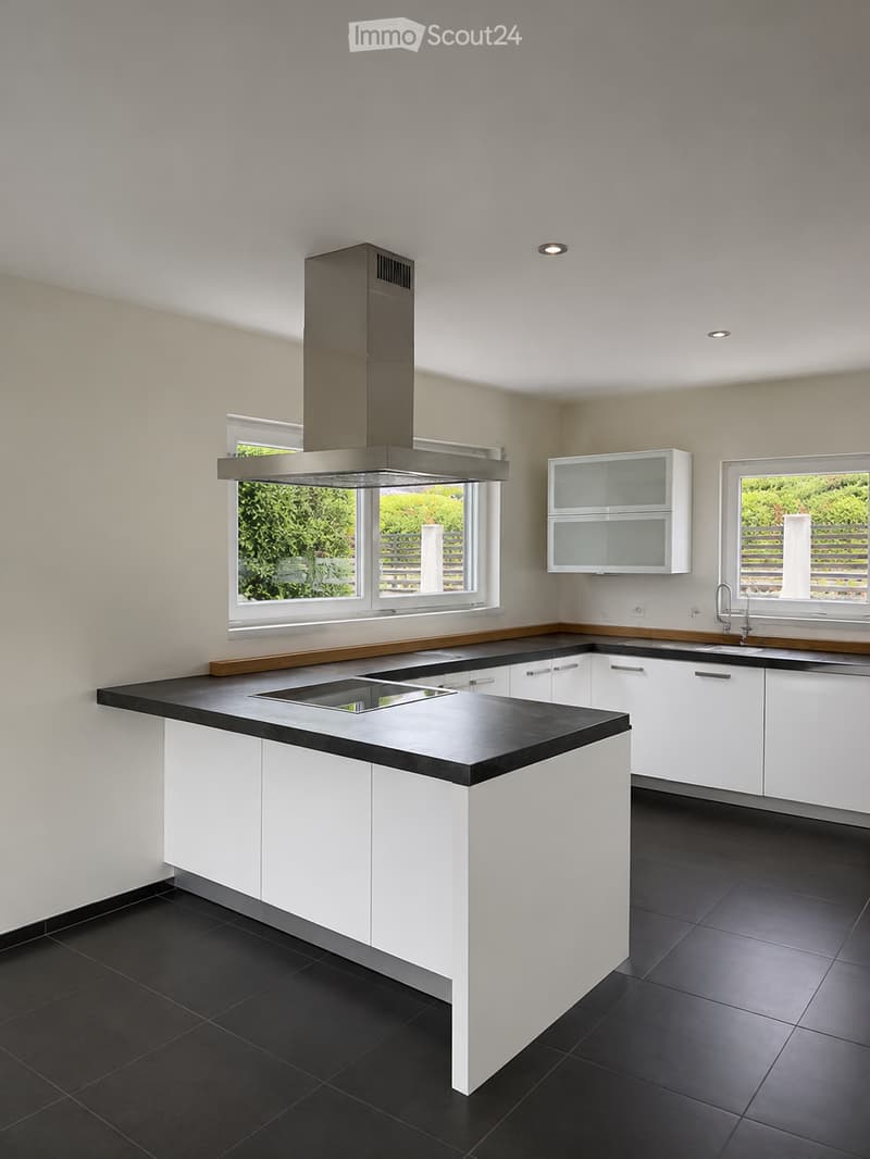 Modern kitchen with white cabinets, black countertops, and a stainless steel range hood. Features large windows with a view of greenery.