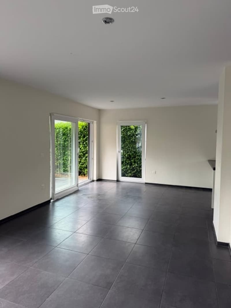 Empty room with dark tiled floor, white walls, and large glass sliding doors leading to a green outdoor area.