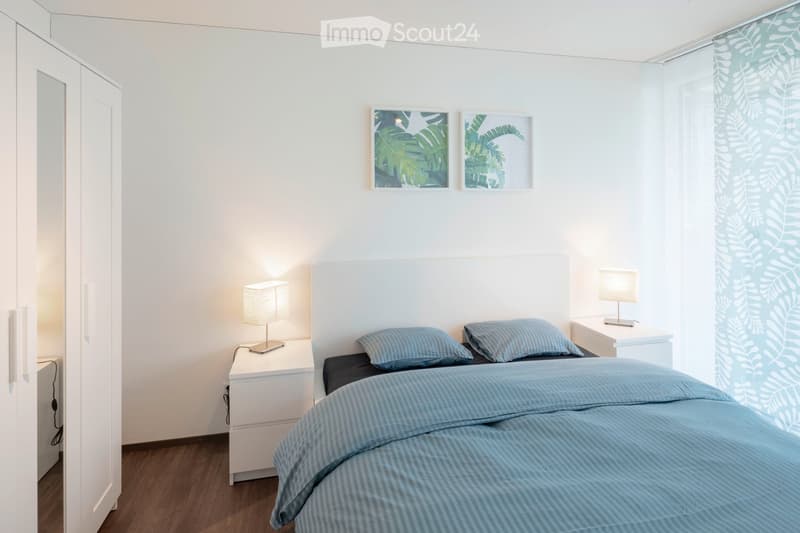 Double bed with blue linens, two nightstands with lamps, wall art with leaf designs, white wardrobe with mirror, dark wooden floor