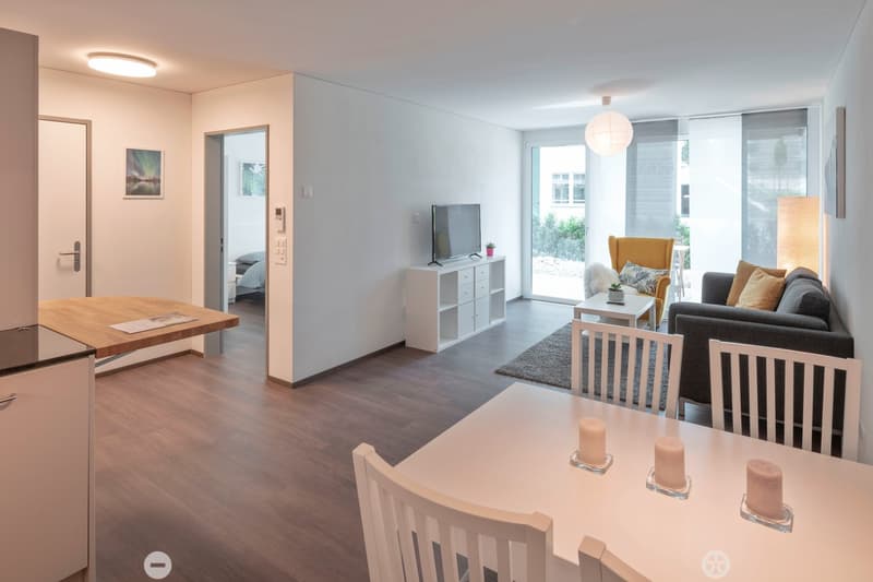Open-plan living area with sofa, coffee table, TV, dining table, dining chairs, lamps, wooden floors, sliding glass doors, open door to bedroom, modern appliances, picture frames, artwork.