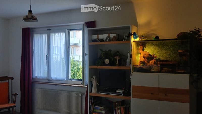A cozy living room with a large window, a wooden chair, a shelf with books and decorative items, a fish tank, and a table with a TV and other items.