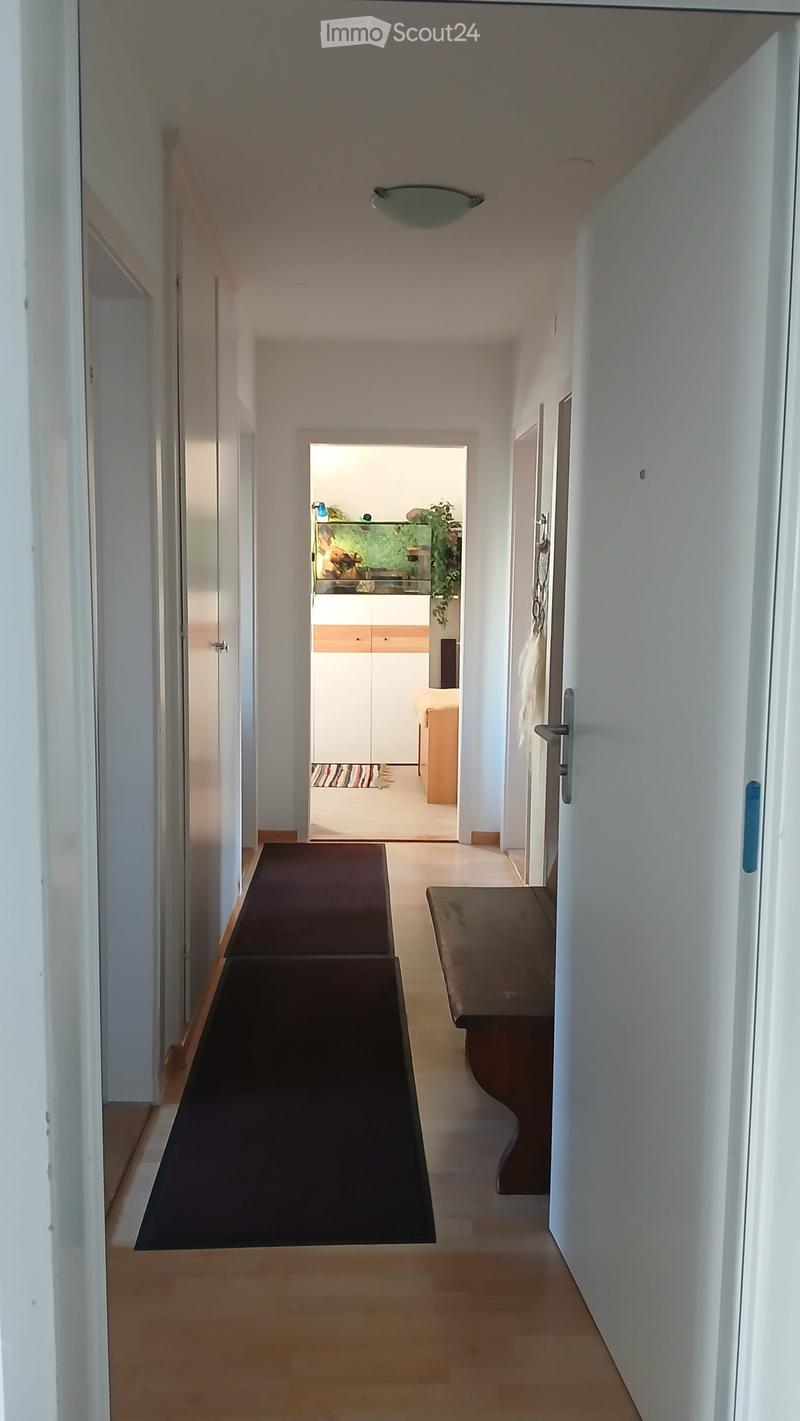 A modern hallway with light-colored wooden flooring, white walls, and a dark runner. A small bench is placed on the right side, and a doorway leads to another room with visible furniture and decor.