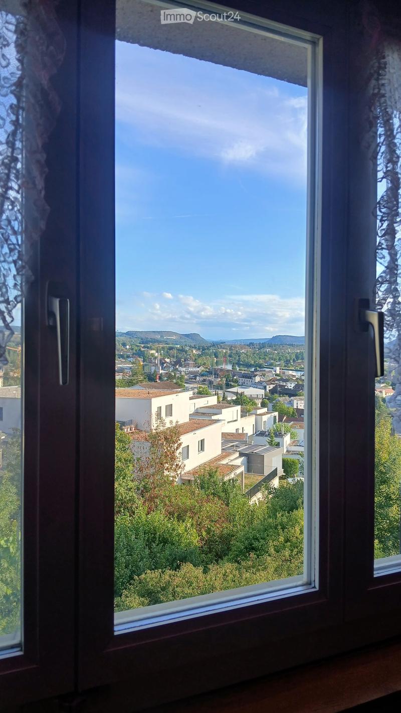 View from a window showing a town with buildings, trees, and distant hills under a clear blue sky.