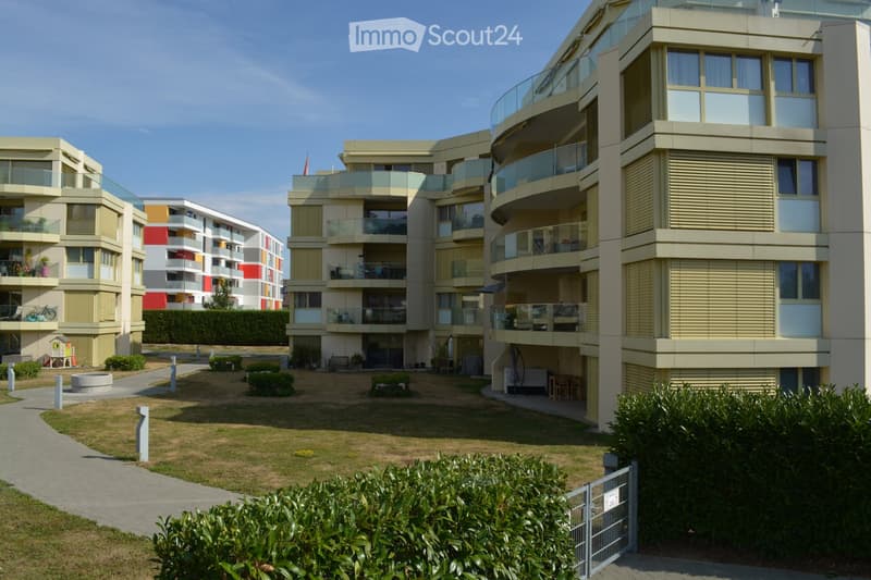 Modern multi-story residential building with balconies, glass railings, and a landscaped courtyard area with pathways and greenery.
