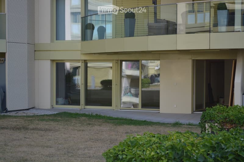 Modern apartment building with large glass sliding doors, a balcony with outdoor furniture, and a small garden area with grass and plants.
