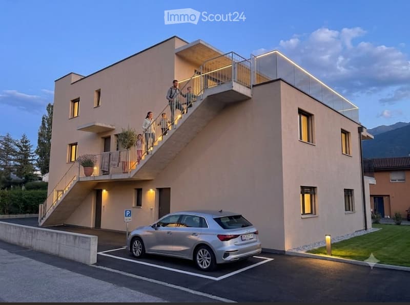 Modern two-story building with a raised ground floor, balcony, and parking space for a car.