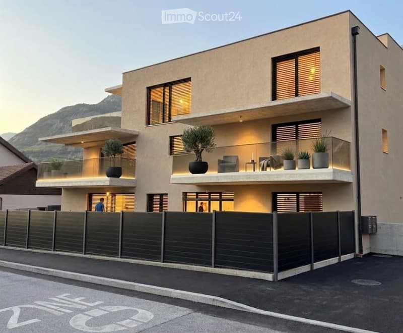 Modern two-story house with large windows, balconies, and potted plants. The building features a sleek design with a contemporary facade and is situated in a scenic area with a mountain view.