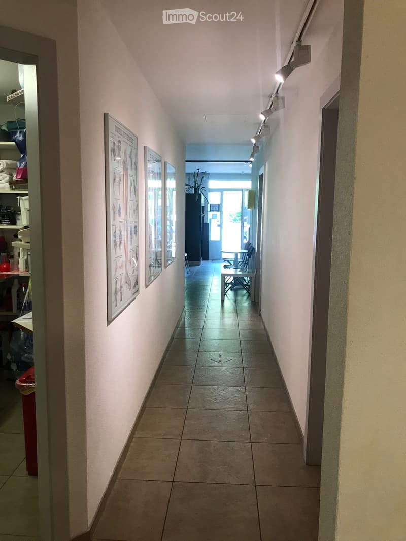 A narrow hallway with tiled flooring and white walls, featuring framed posters on one side and a small seating area visible at the end.