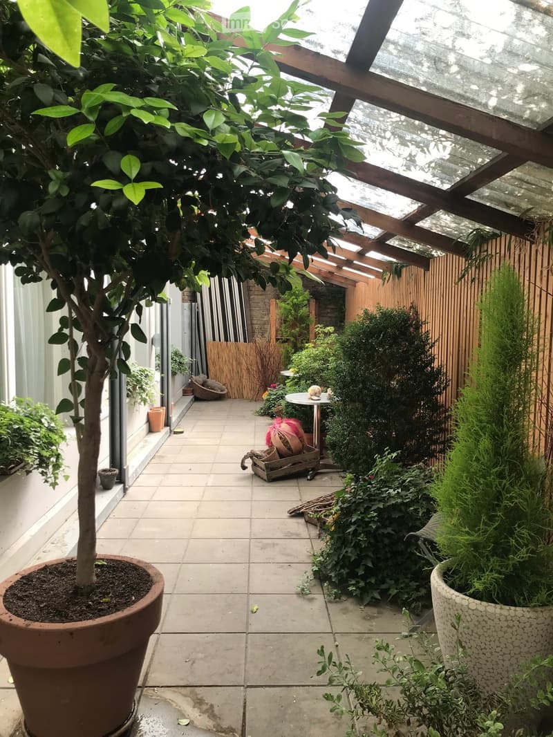 A covered outdoor patio with a tiled floor, potted plants, and a small table with decorative items. The area is enclosed by wooden fencing and has a pergola-style roof allowing natural light.