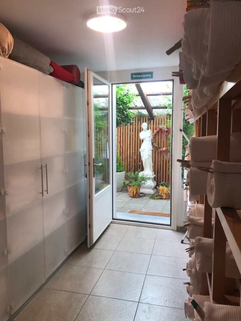 Hallway with tiled floor, large open sliding doors leading to a garden area, shelves with neatly stacked towels, and a white statue visible in the garden.