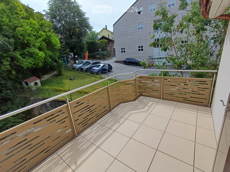 Balcony with beige tiled floor and wooden balustrade, overlooking a green lawn, parked cars, and neighboring buildings.